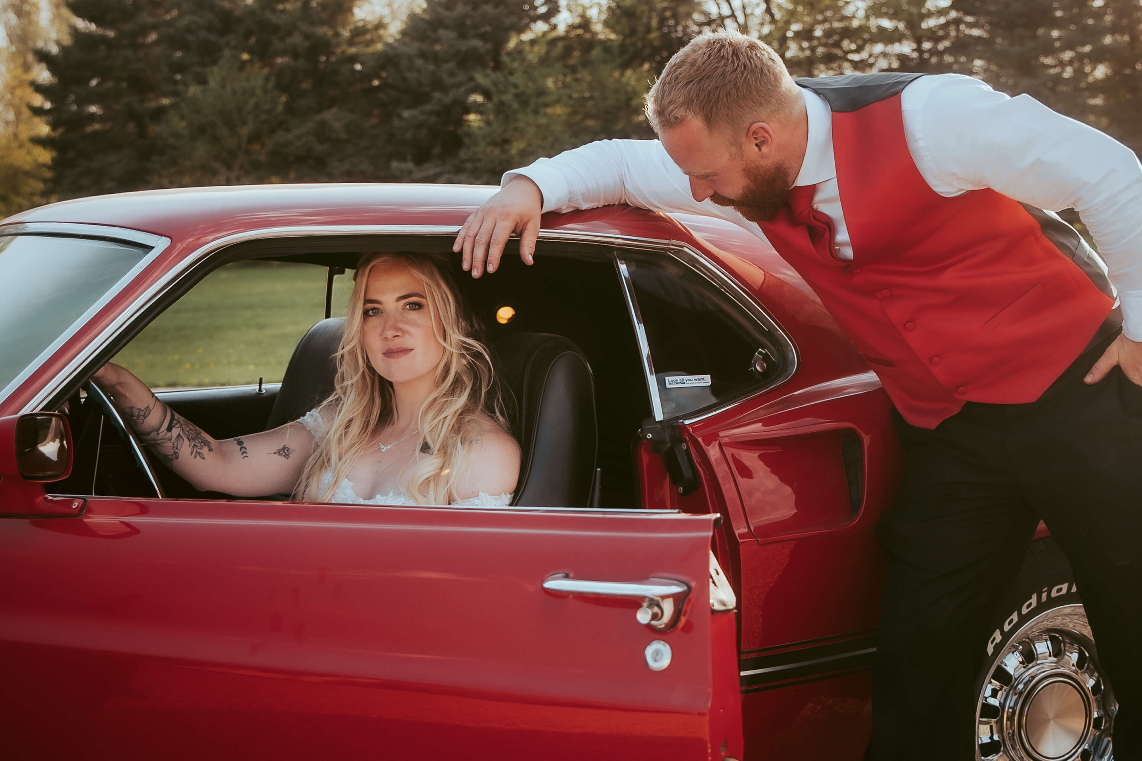 Bride in a white dress sitting in a bright red ford mustang class muscle car with the groom leaning over the card in a red suit and black pants.